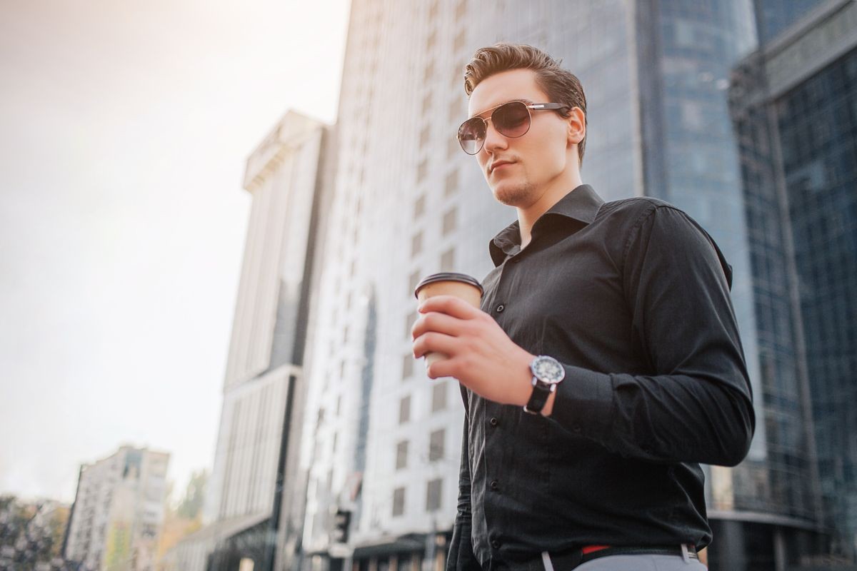 Confident young man stand in city and pose. He hold cup of drink and look forward. He stand in front of building. He looks like a buisnessman.