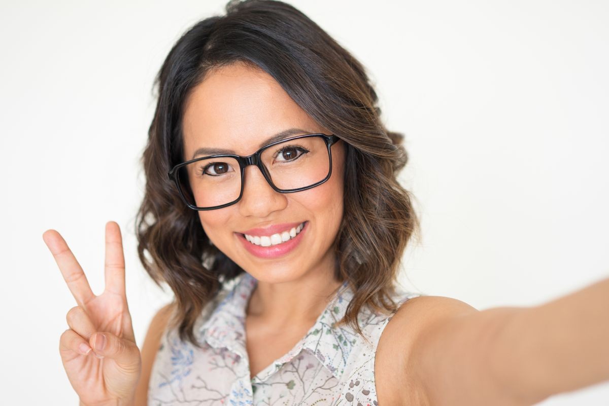 Closeup portrait of smiling young beautiful dark-haired woman looking at camera, taking selfie photo and showing victory sign. Selfie photo concept. Isolated front view on white background.