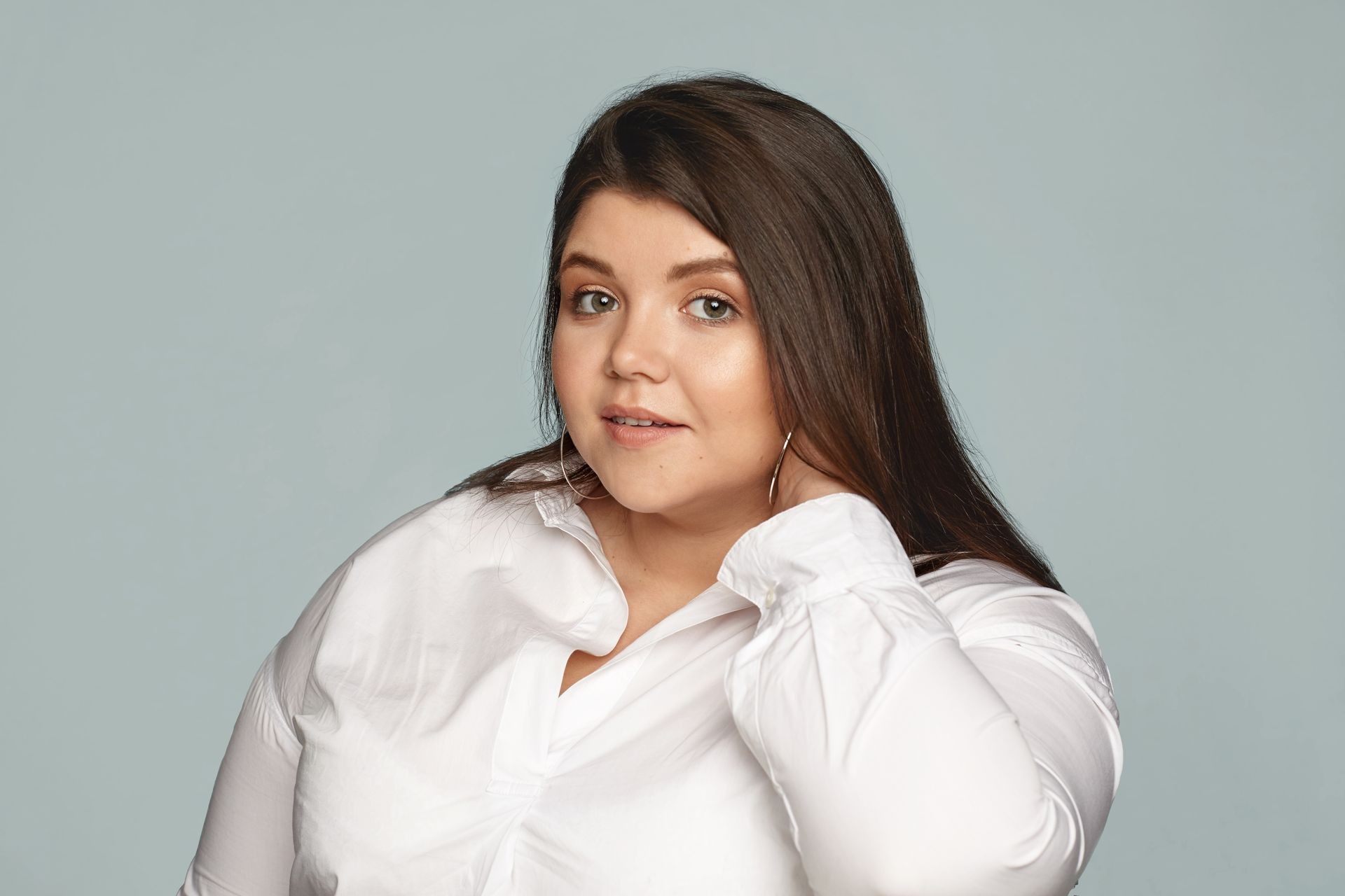 Picture of stylish young overweight female employee wearing white shirt and large round earrings touching her neck. Neat beautiful chubby woman posing against gray scopyspace tudio wall background